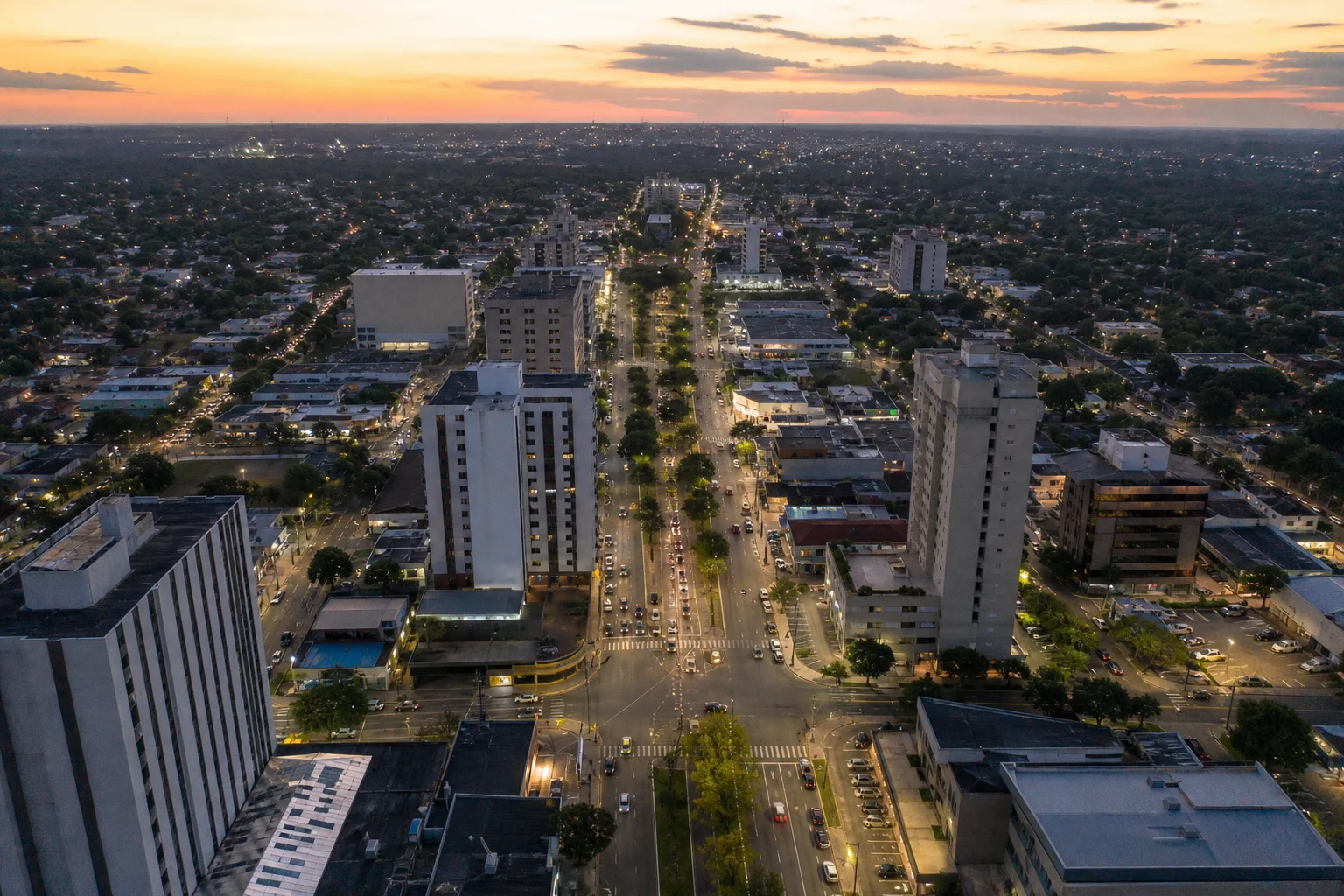 Vista aérea de avenida urbana ao entardecer, com prédios comerciais e o traçado da cidade se estendendo ao fundo.